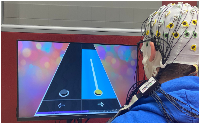 This picture represents the new visual interface of the Brain Hero visual BCI training environment, together with a user actually using it. The game visual looks like notes on a guitar FretBoard, each note representing a specific mental task for the BCI.